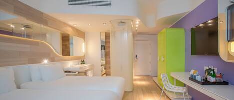 A modern hotel room with a bed, a bathroom, a small table with toiletries, and a mirror.