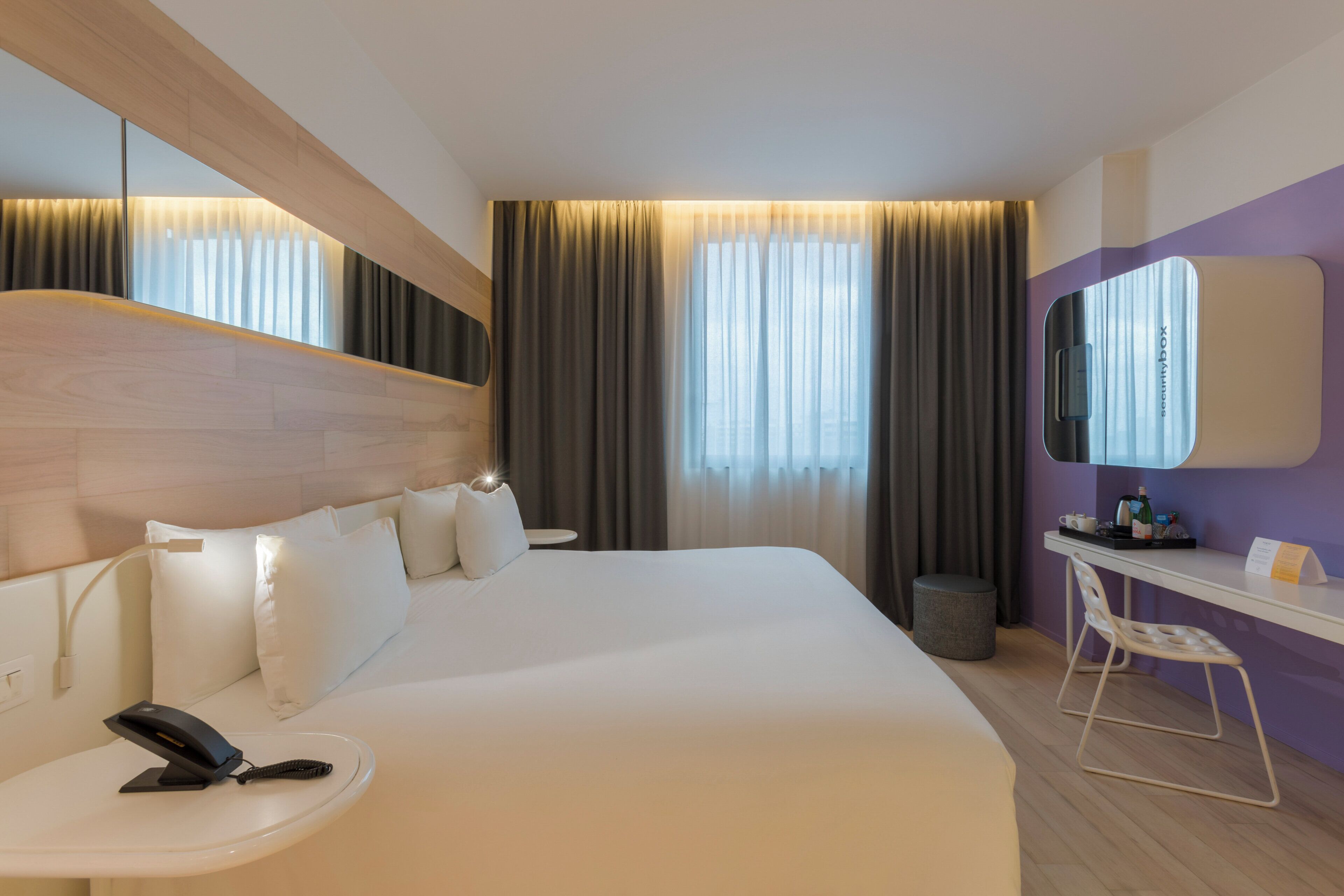 Une chambre d’hôtel avec un grand lit, un bureau, une chaise, un miroir et une fenêtre avec des rideaux.