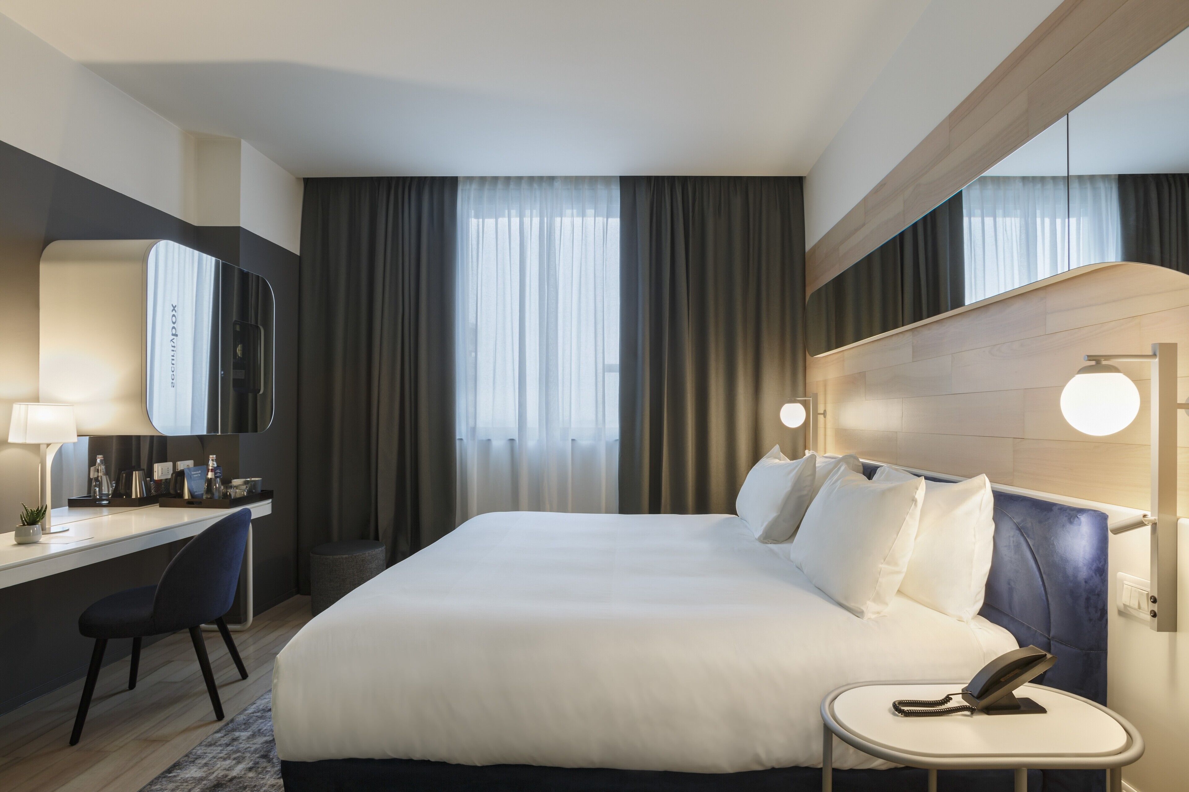 Une chambre d’hôtel moderne dotée d’un grand lit, d’un bureau avec une chaise, d’un miroir et d’une table de chevet avec un téléphone.