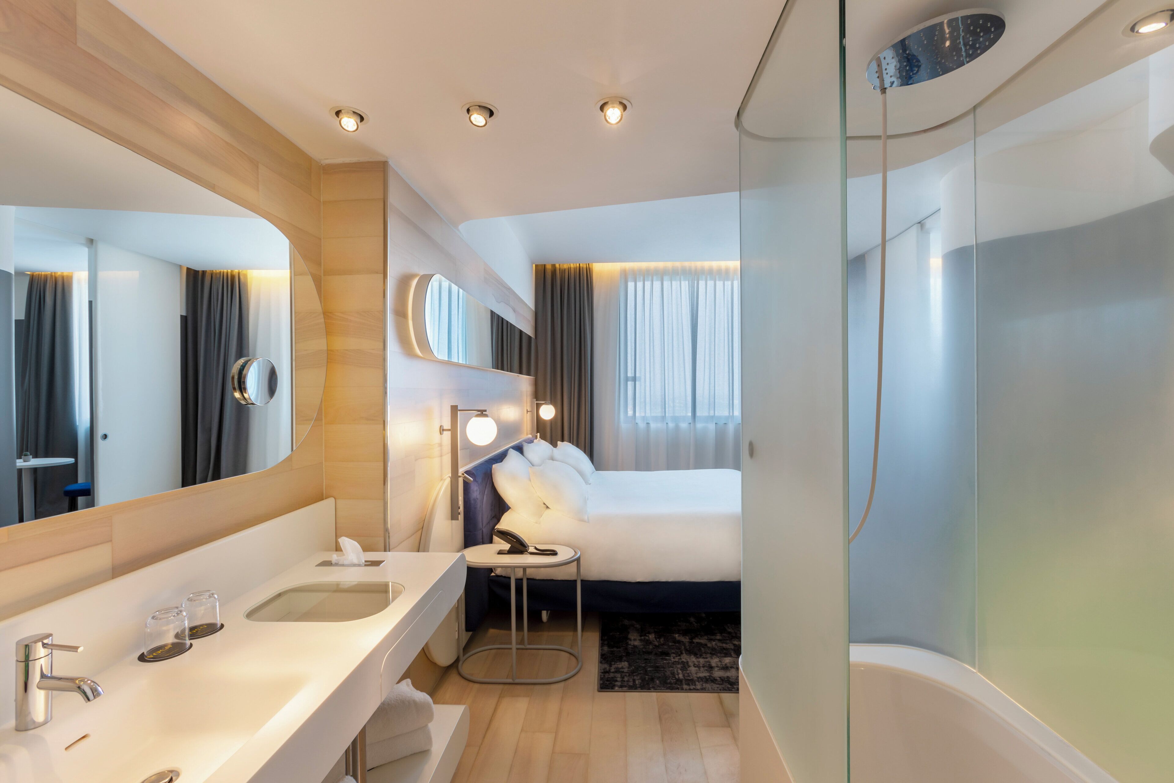 Een moderne hotelbadkamer met een grote spiegel, een ligbad, een rond nachtkastje en een aparte wastafel.