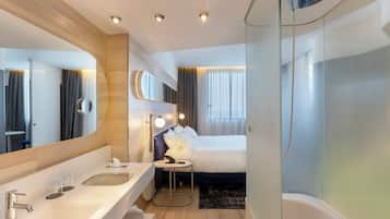 Een moderne hotelbadkamer met een grote spiegel, een ligbad, een rond nachtkastje en een aparte wastafel.