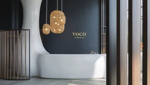 A modern hotel reception area with a curved reception desk, a large black wall with the hotel name, and two hanging wicker pendant lights.