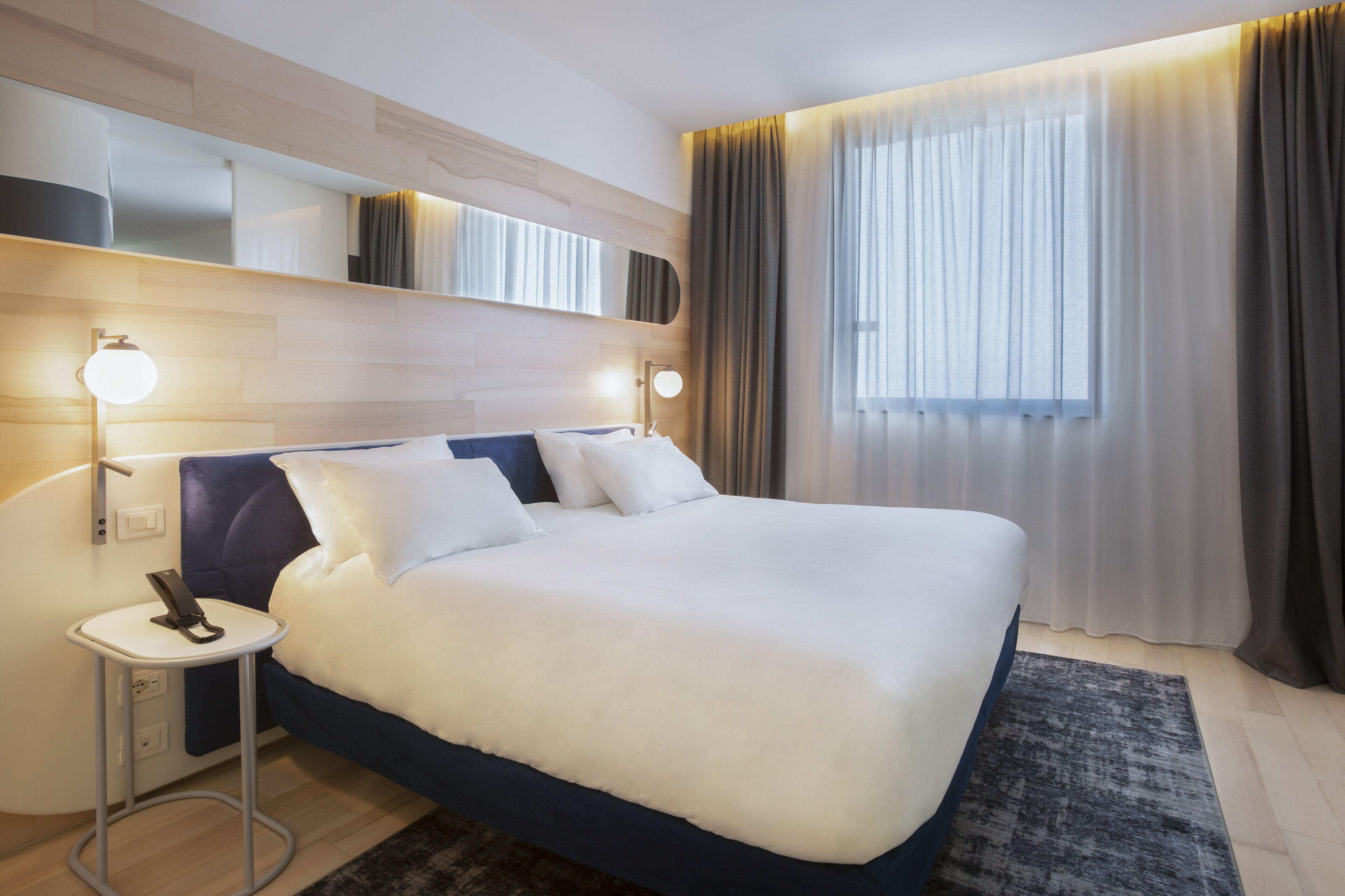 Une chambre d’hôtel moderne avec un grand lit, des lampes de chevet, une petite table et une grande fenêtre avec des rideaux.