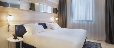 A modern hotel room with a large bed, bedside lamps, a small table, and a large window with curtains.
