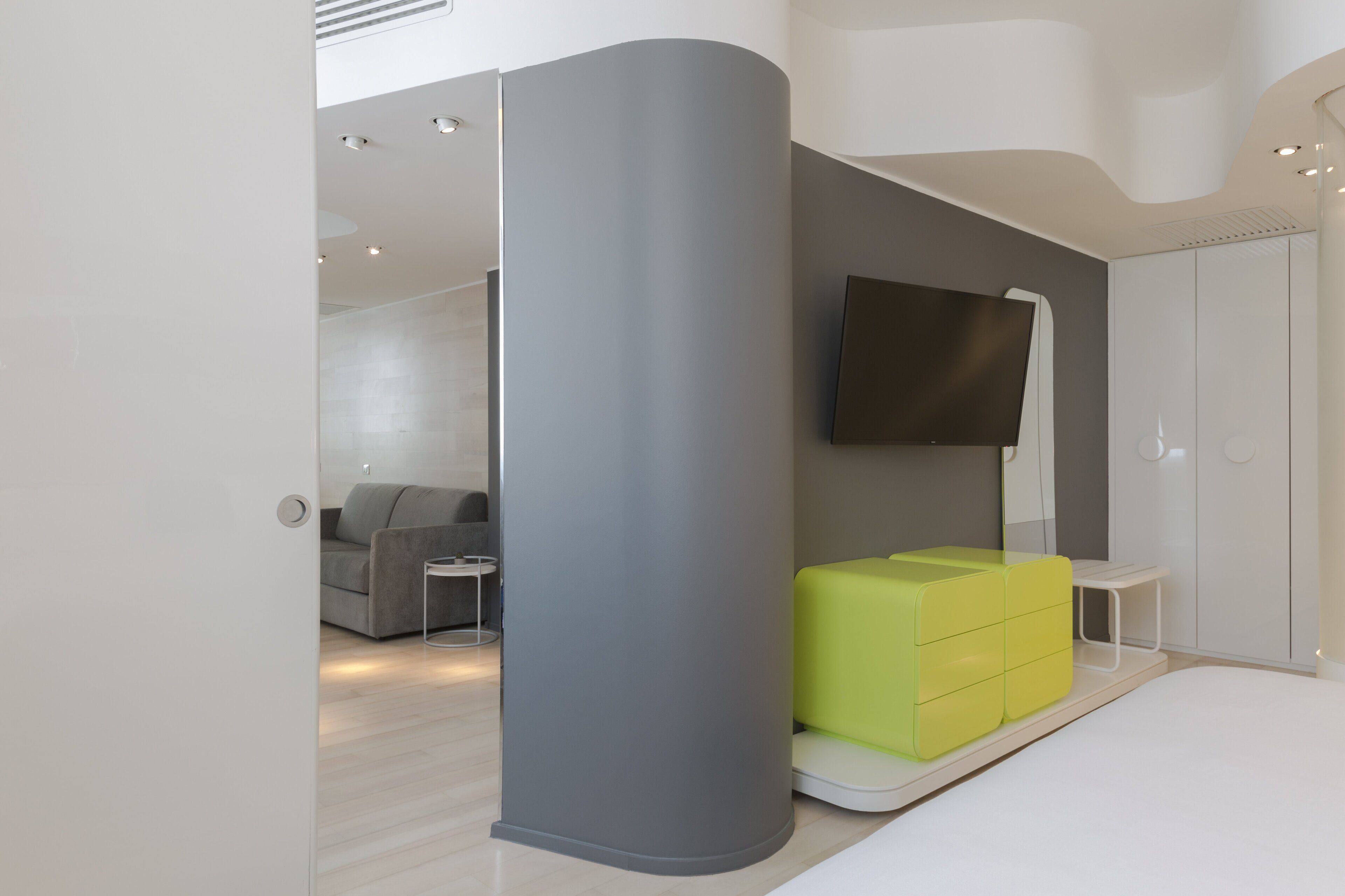 A modern room with a grey pillar, a green sofa, a flat-screen TV, and a white bed.