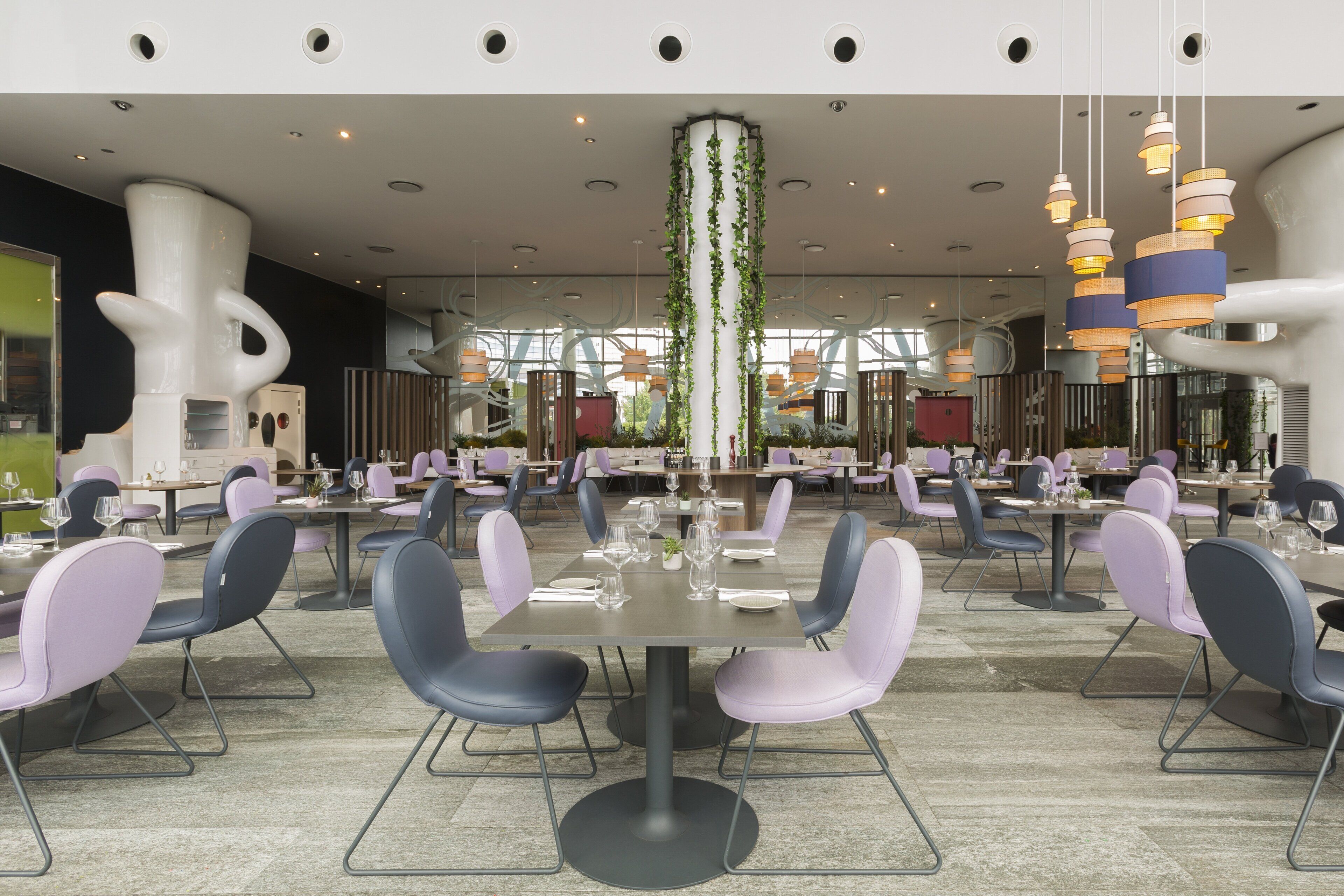 Een moderne eetruimte met ronde tafels en stoelen, een centrale kolom met beplanting en hangende hanglampen.