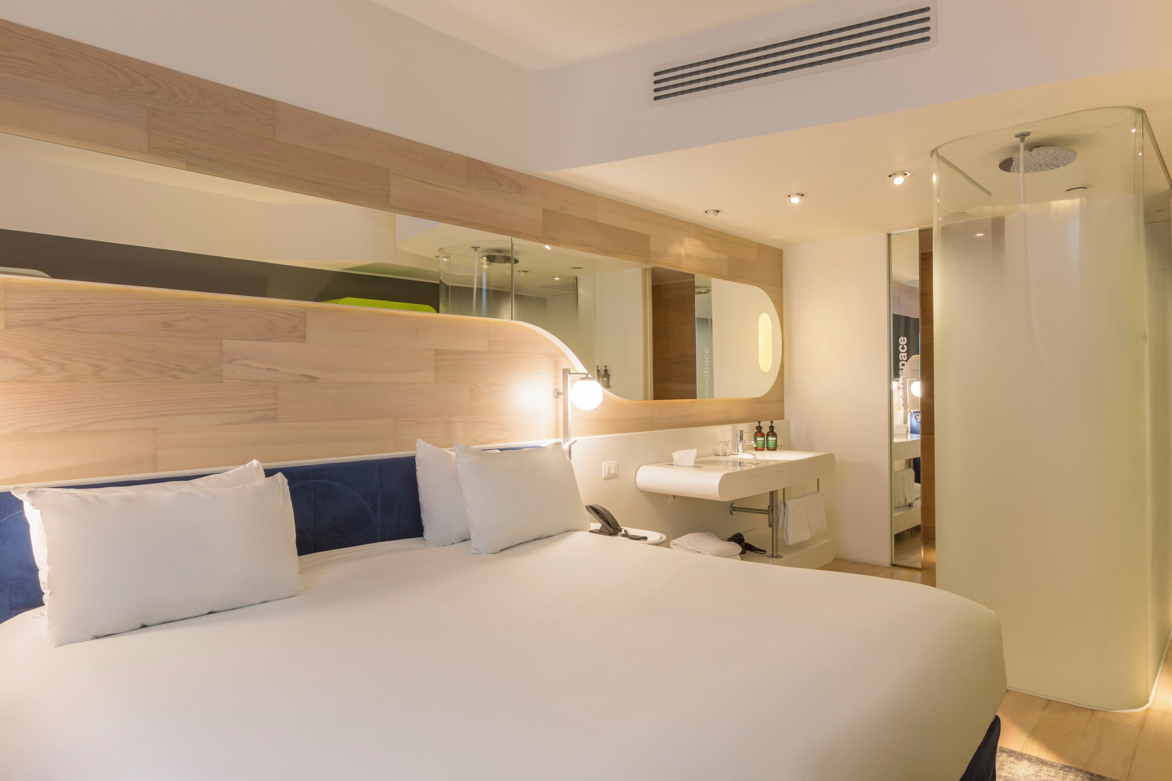 A modern hotel room with a large bed, a bedside table, a mirror, a bathroom with a sink, and a shower.