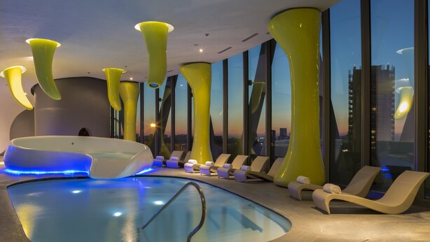 A modern indoor pool area with a hot tub, lounge chairs, and large windows offering a city view.