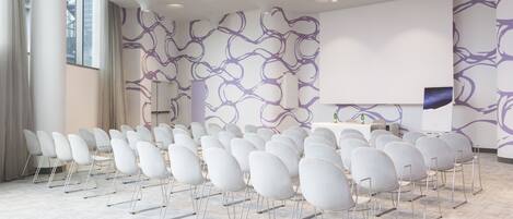 Un salón de conferencias con filas de sillas blancas, una pizarra grande y arte abstracto morado en las paredes.