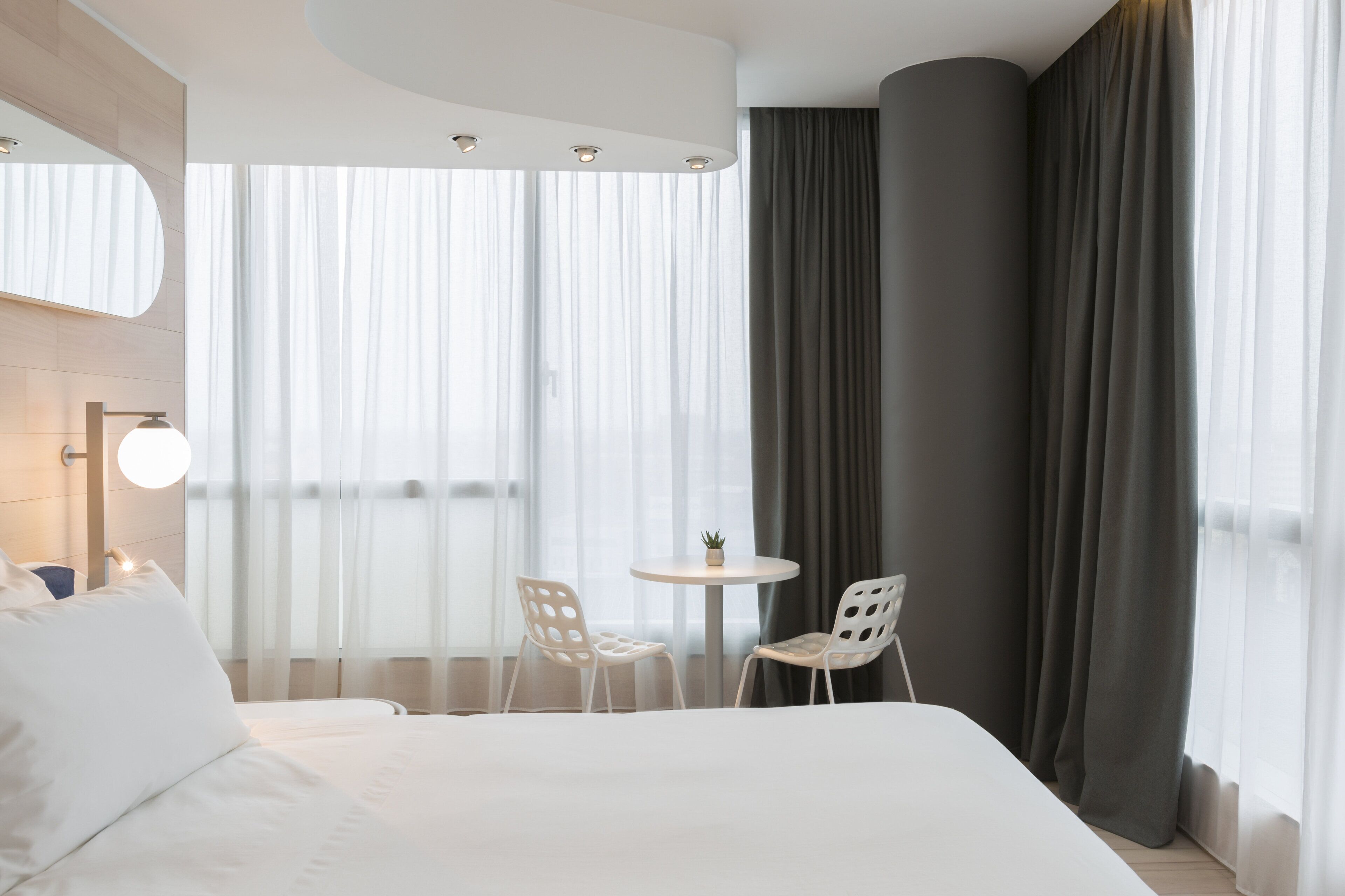 A hotel room with a large bed, two white chairs, a small round table with a plant, and a large window with sheer curtains.
