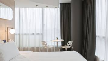 A hotel room with a large bed, two white chairs, a small round table with a plant, and a large window with sheer curtains.
