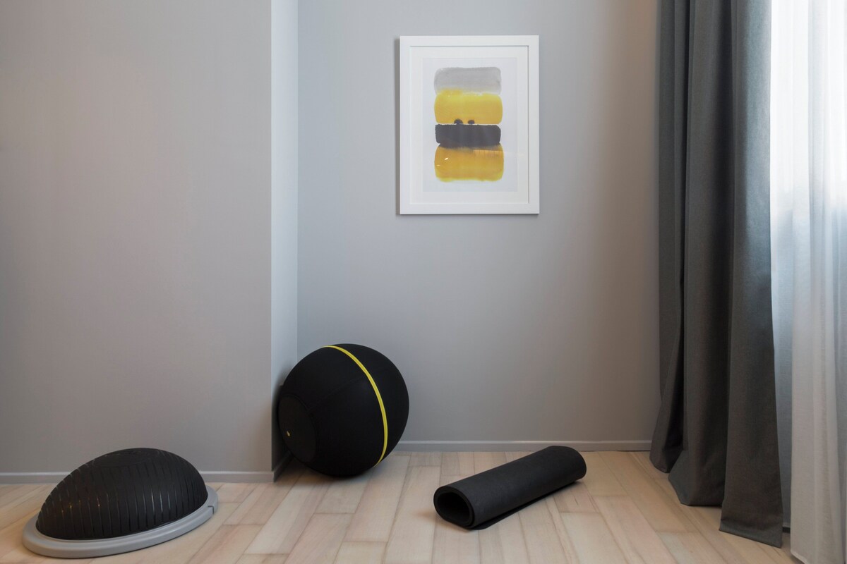 A room with a grey wall, a framed abstract painting, a black exercise ball, and a yoga mat.