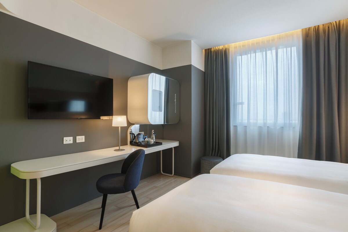 A hotel room with a bed, a desk with a chair, a TV mounted on the wall, and a window with curtains.