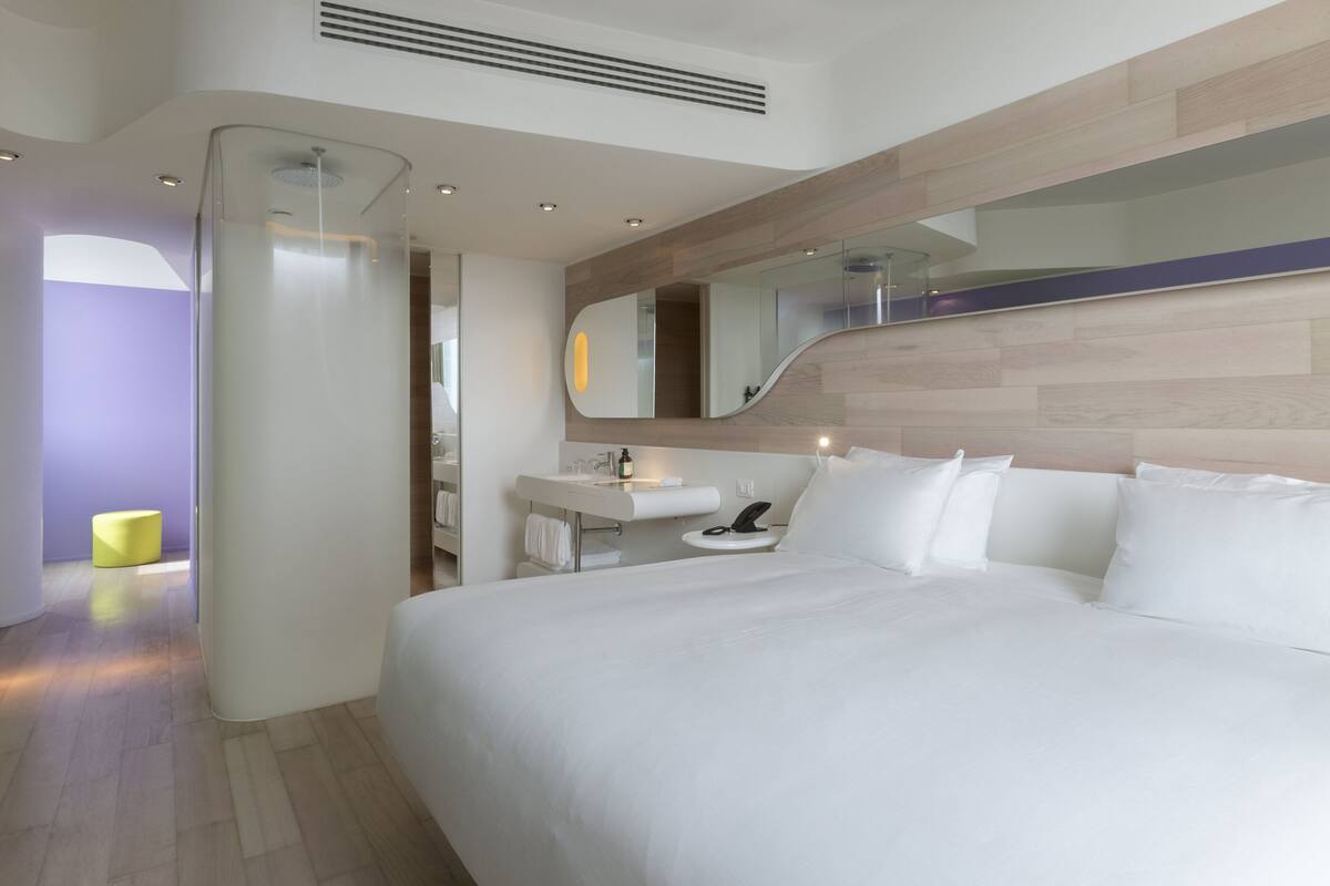 A modern hotel room with a large bed, a bedside table, a mirror, and a bathroom with a sink and toilet.