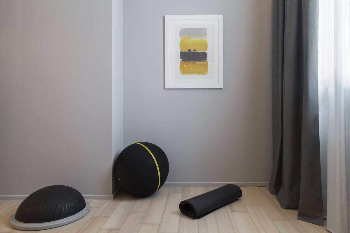 A room with a grey wall, a framed abstract painting, a black exercise ball, and a yoga mat.
