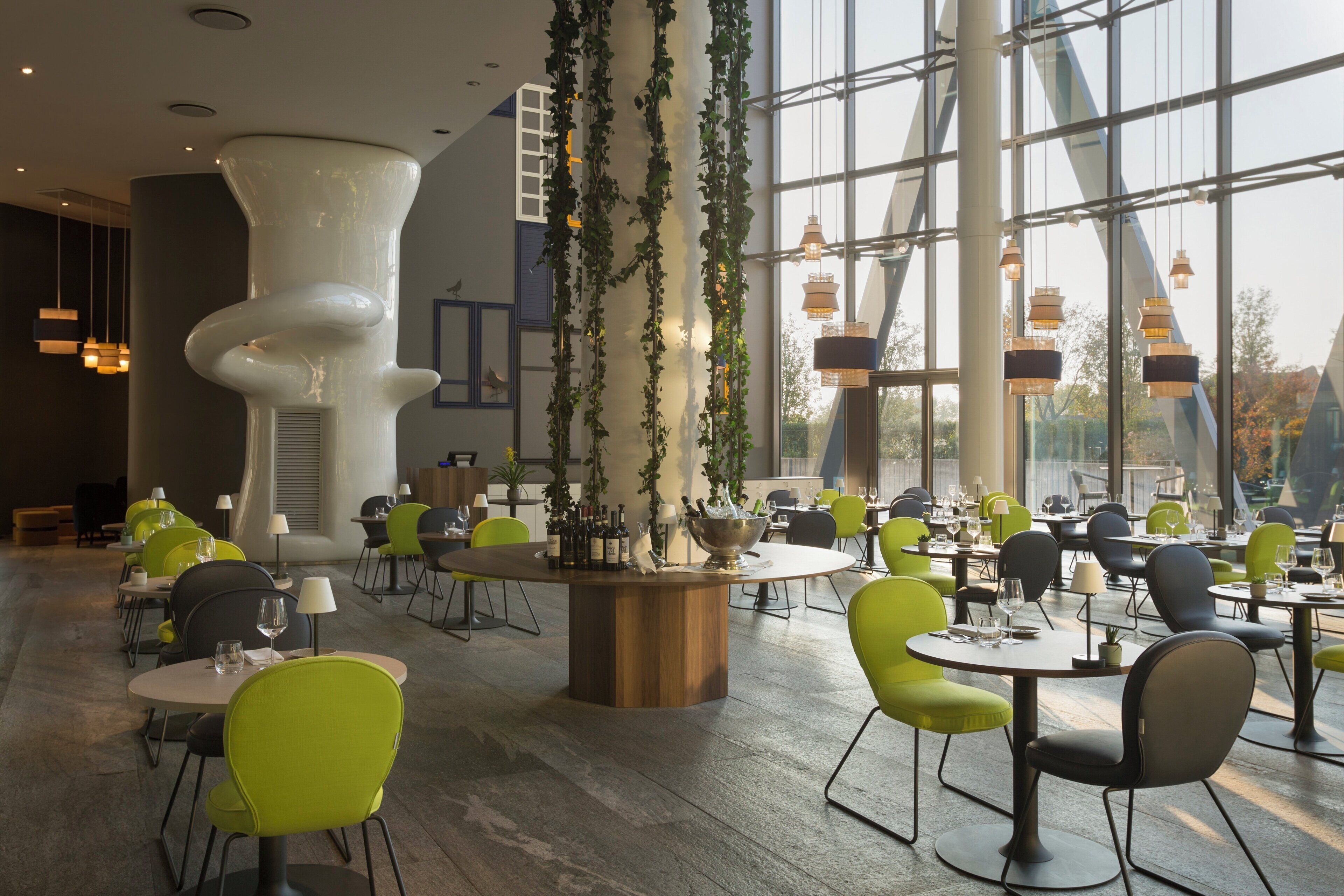 Un espace repas moderne avec des tables et des chaises rondes, un bar central où sont exposées des bouteilles, et de grandes fenêtres qui laissent entrer la lumière naturelle.