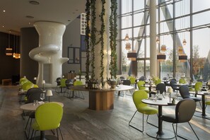 A modern dining area with round tables and chairs, a central bar area with bottles, and large windows allowing natural light. - voco Milan - Fiere by IHG (Milan)