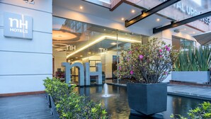L’entrée d’un hôtel, avec une façade moderne en verre, une fontaine à l’avant et une enseigne indiquant « nh hotels ».