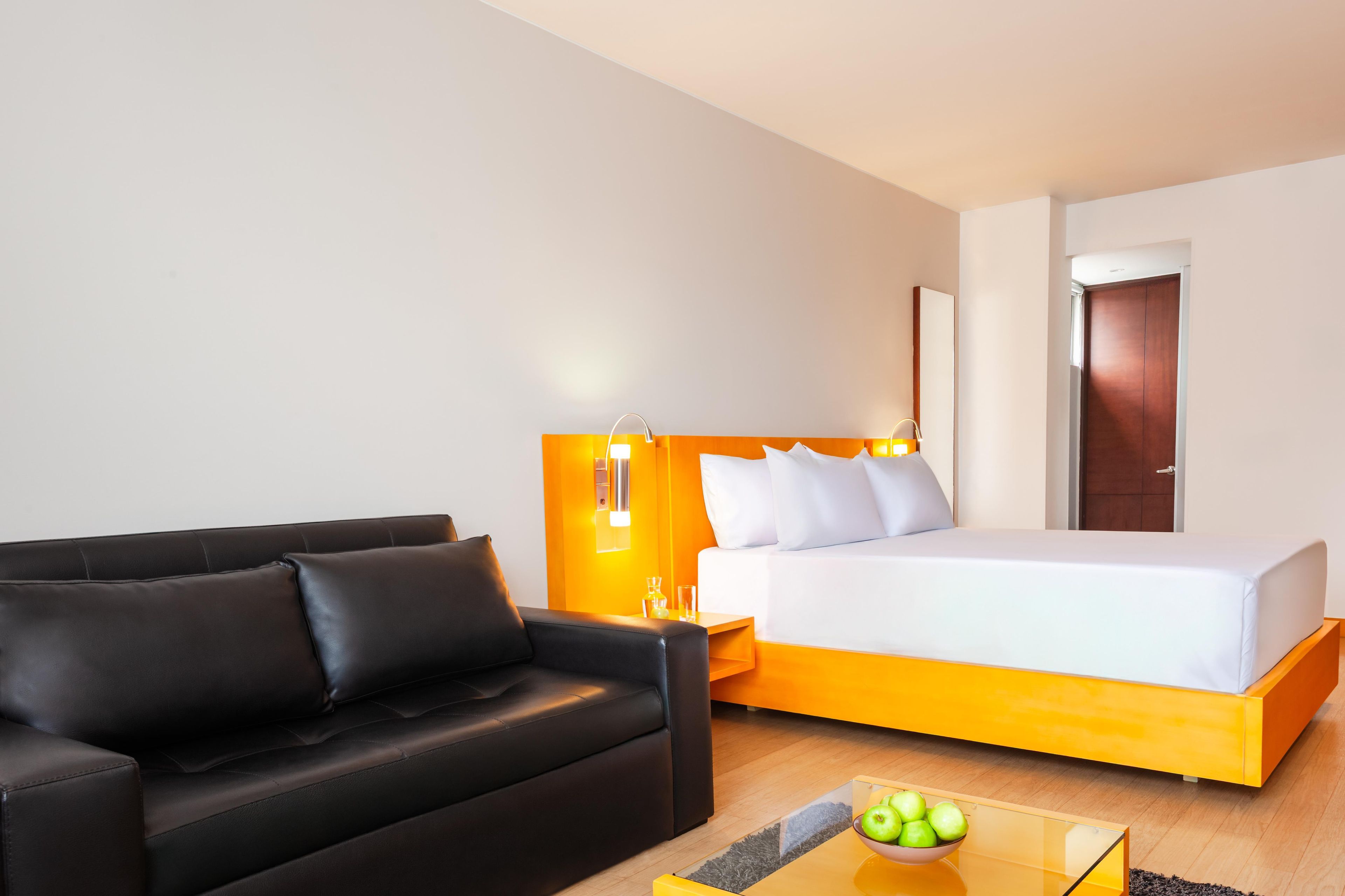 A hotel room with a black leather sofa, a bed with white bedding, and a glass coffee table with a bowl of apples.