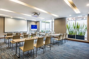 A conference room with tables, chairs, a projector, and a large window with plants.