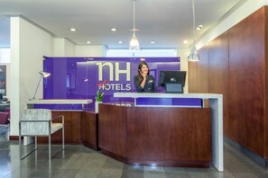 Un comptoir d’accueil au design moderne, une grande enseigne indiquant « NH Hotels », et un espace détente avec un fauteuil.