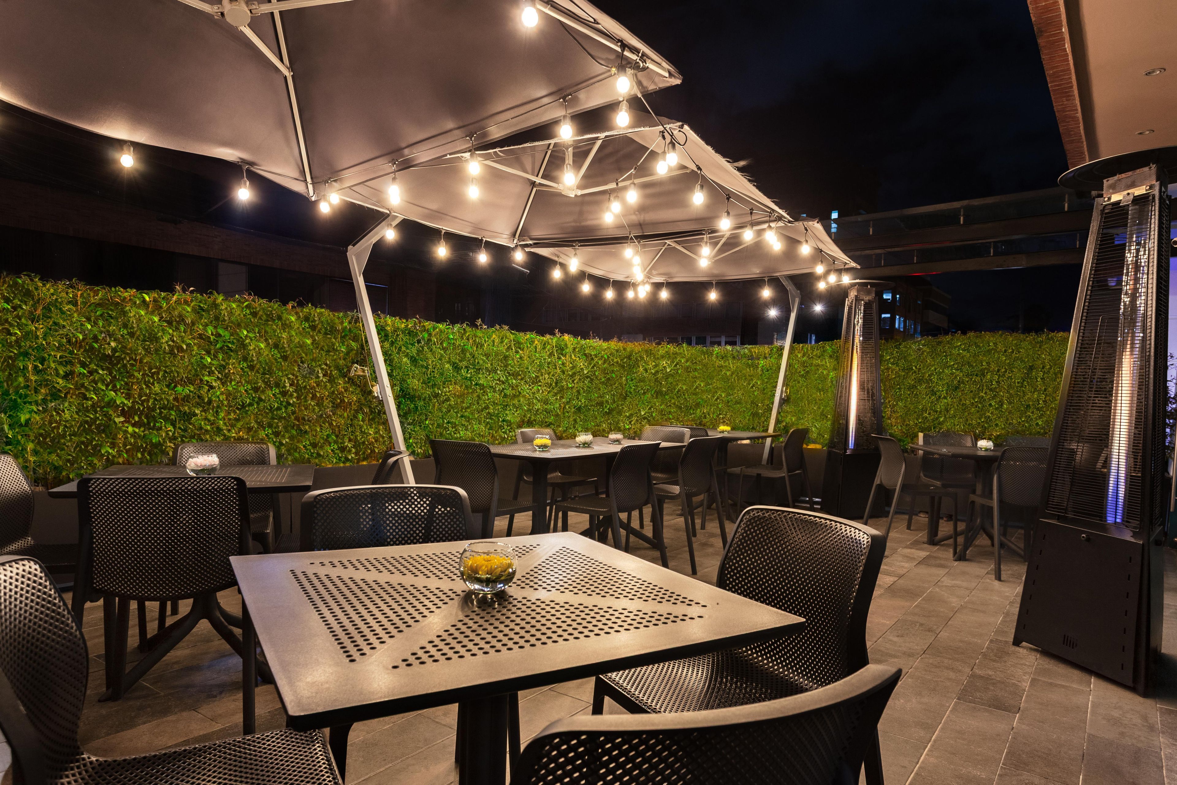Un espace de restauration en plein air, avec des tables et des chaises, abrité par une tonnelle décorée de guirlandes lumineuses, et un mur végétal en arrière-plan.