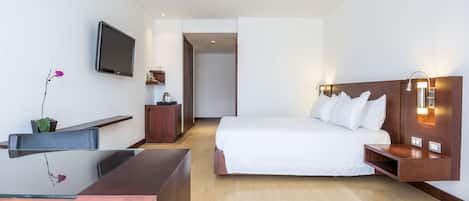 A hotel room with a bed, bedside table, desk, chair, TV, and a small plant.