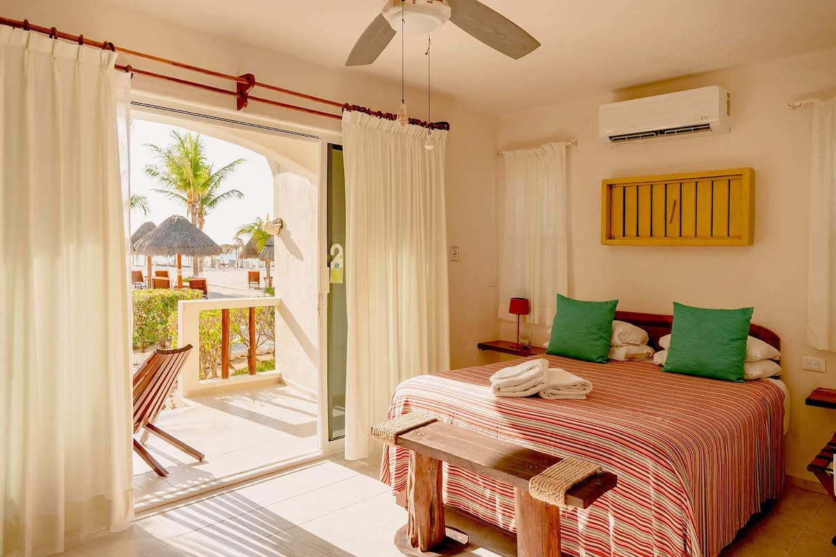 Habitación de hotel con cama, una banca, una mesita de noche, una lámpara roja y vista a la playa con palmeras.