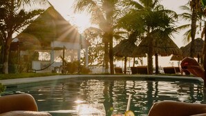 A poolside view with a glass of iced drink and a martini glass, surrounded by palm trees and a thatched-roof structure.