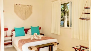 A bedroom with a bed, bedside tables, a hanging macrame wall hanging, a window with curtains, and a small wooden table.