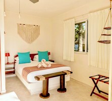 A bedroom with a bed, bedside tables, a hanging macrame wall hanging, a window with curtains, and a small wooden table.