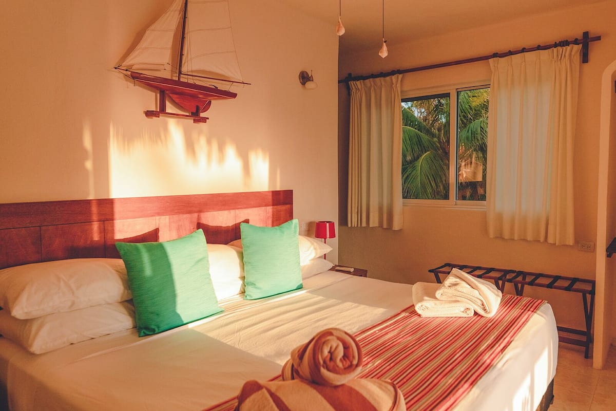 Habitación de hotel con cama, cabecera de madera, decoración de velero, ventana con cortinas y vistas a palmeras.