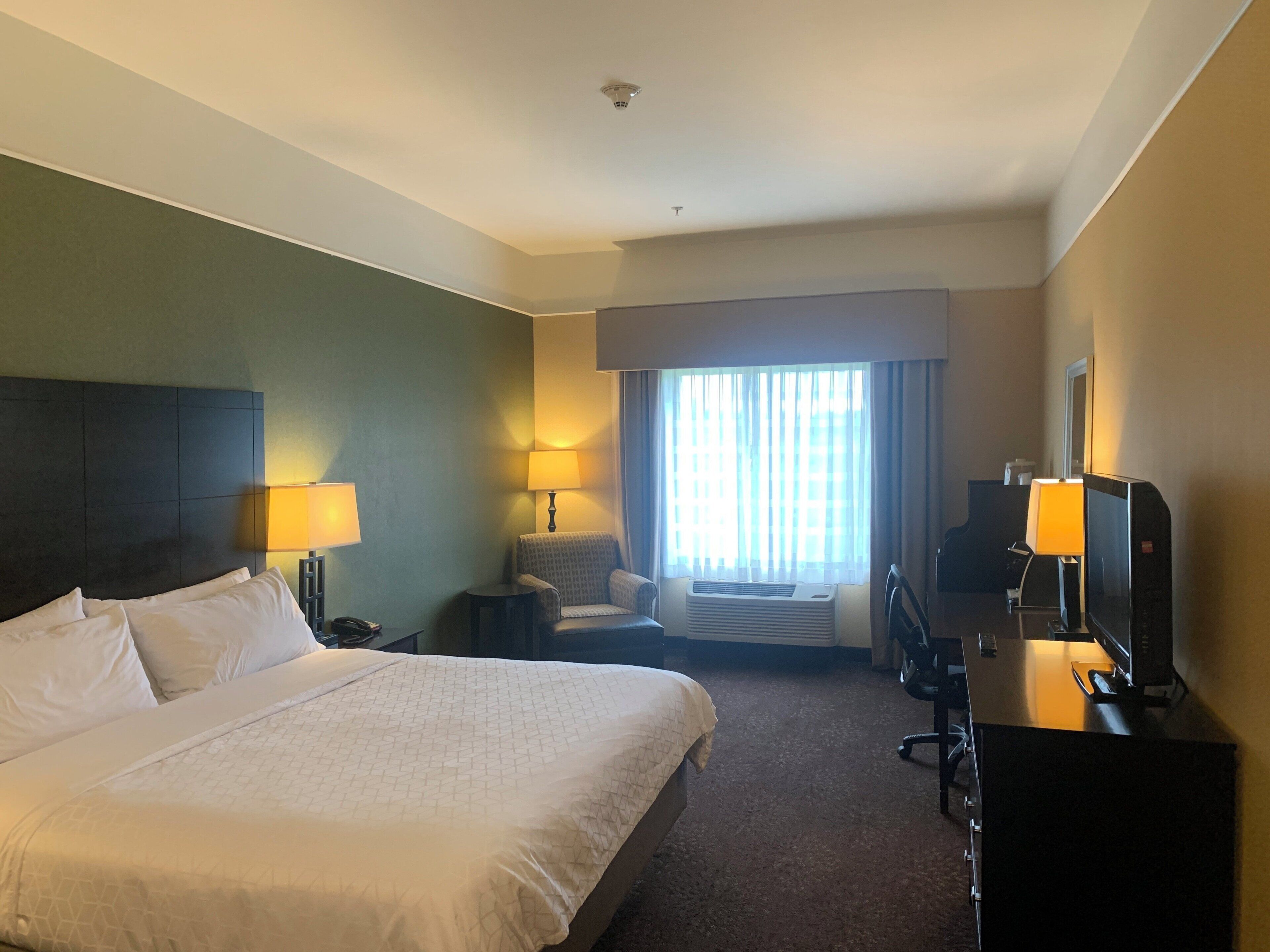 Habitación de hotel con una cama grande, dos mesitas de noche con lámparas, una silla, un escritorio con computadora y un ventanal con cortinas.