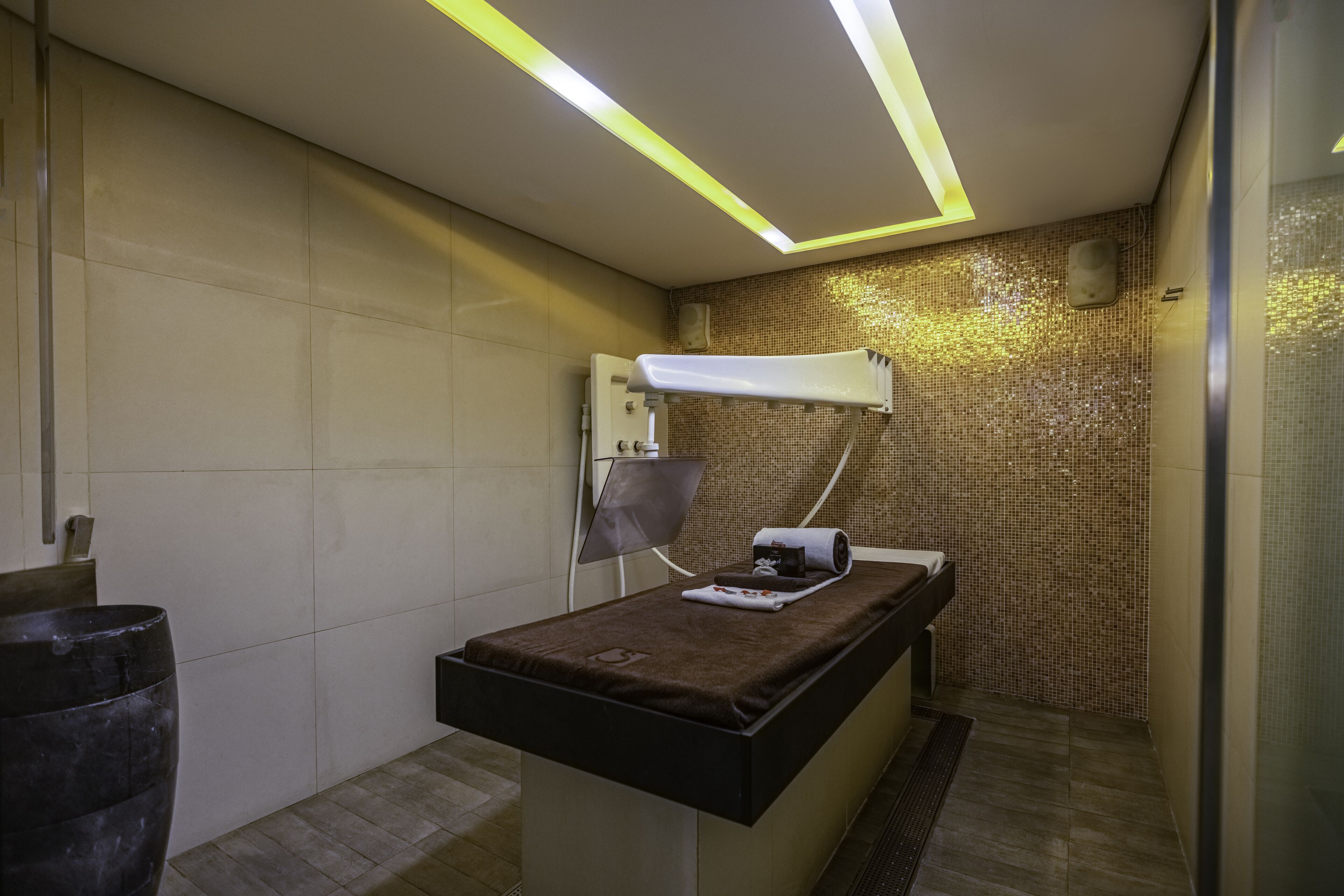 A modern examination room with a medical examination table, a wall-mounted examination light, and a tiled mosaic wall.