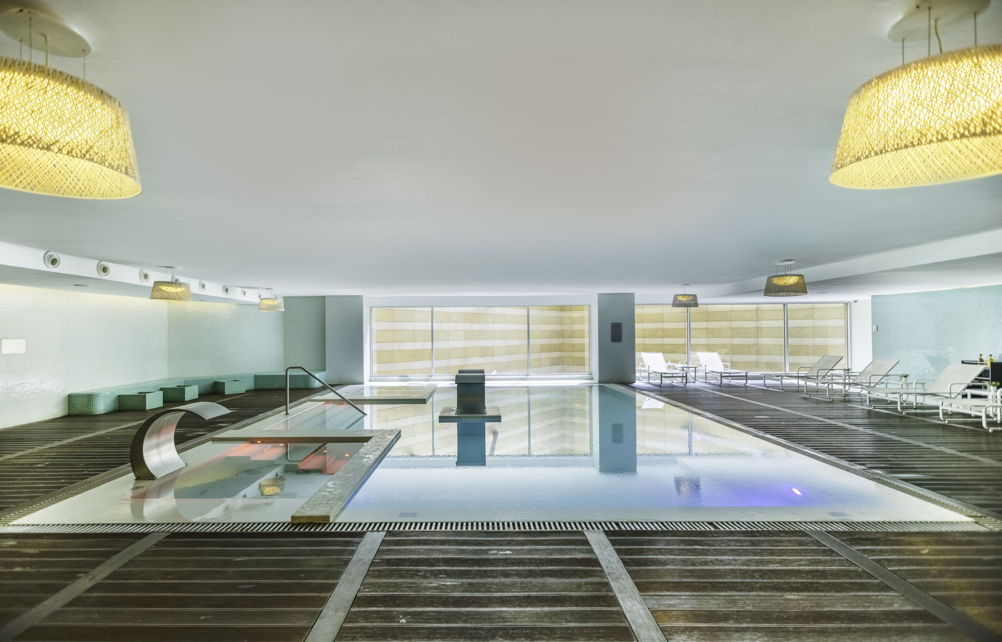 A modern indoor swimming pool with clear glass sides, a central water feature, and lounge chairs along the sides.