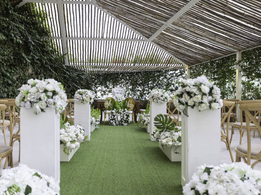 An aisle with white floral arrangements and wicker chairs, leading to a white altar with a green carpet.