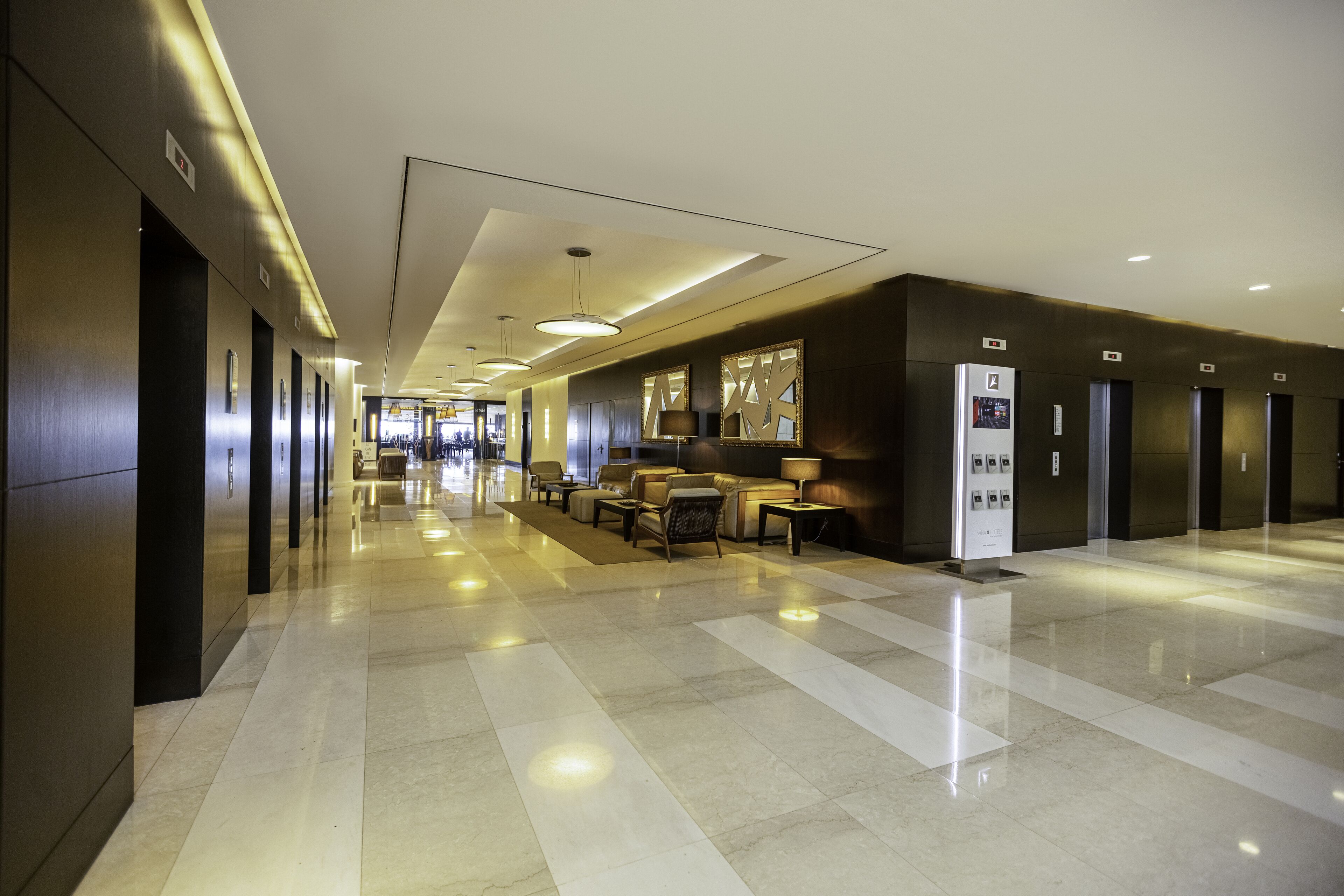 Un hall d’hôtel moderne avec un long couloir, un espace détente avec des tables et des chaises, et un comptoir d’accueil.