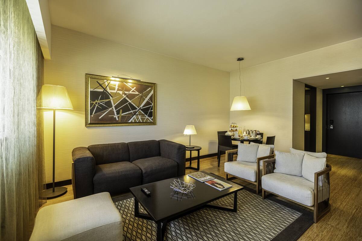 A modern hotel room with a sofa, armchairs, a coffee table, and a dining area.
