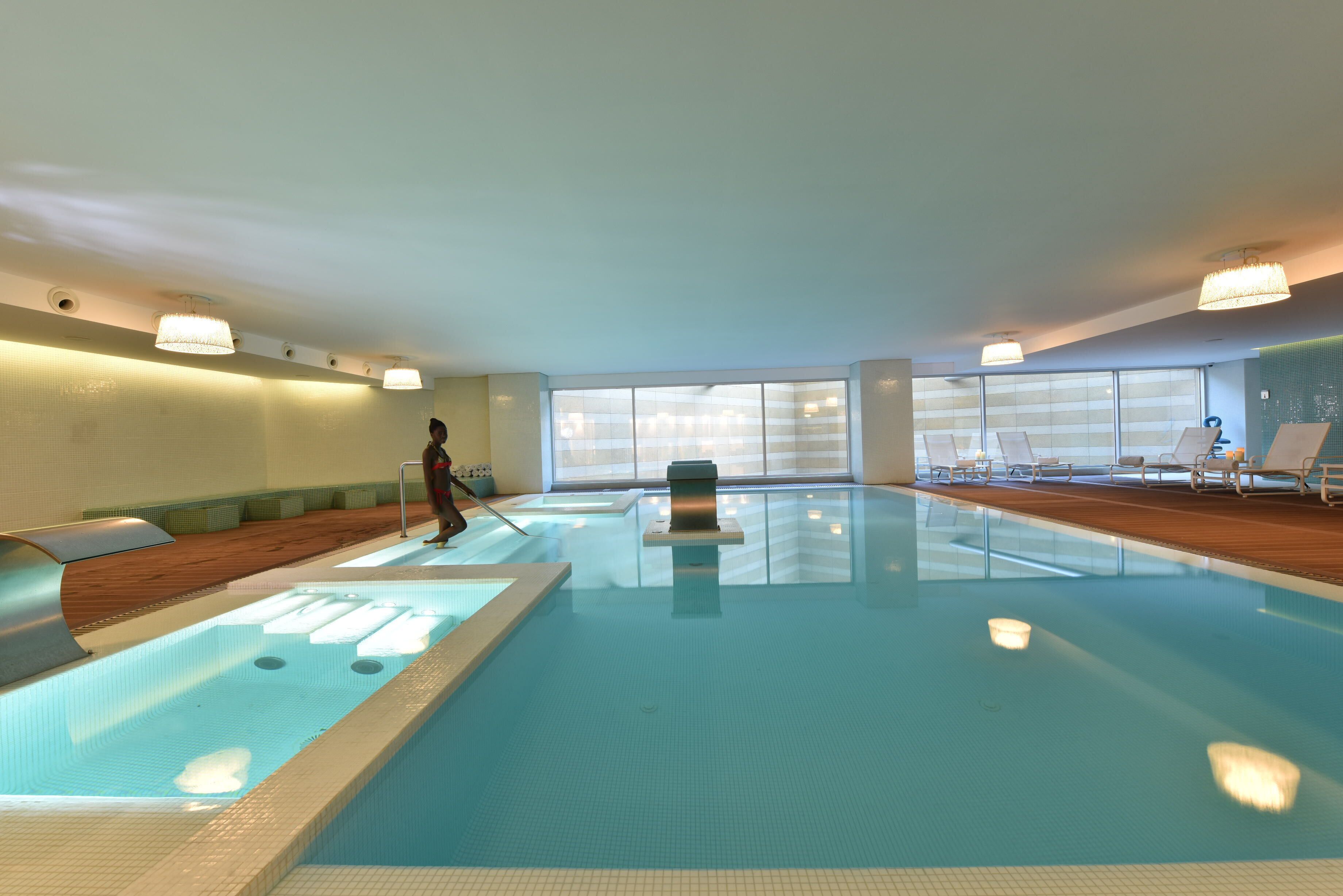 An indoor swimming pool with a clear view of the pool area, lounge chairs, and a person swimming.