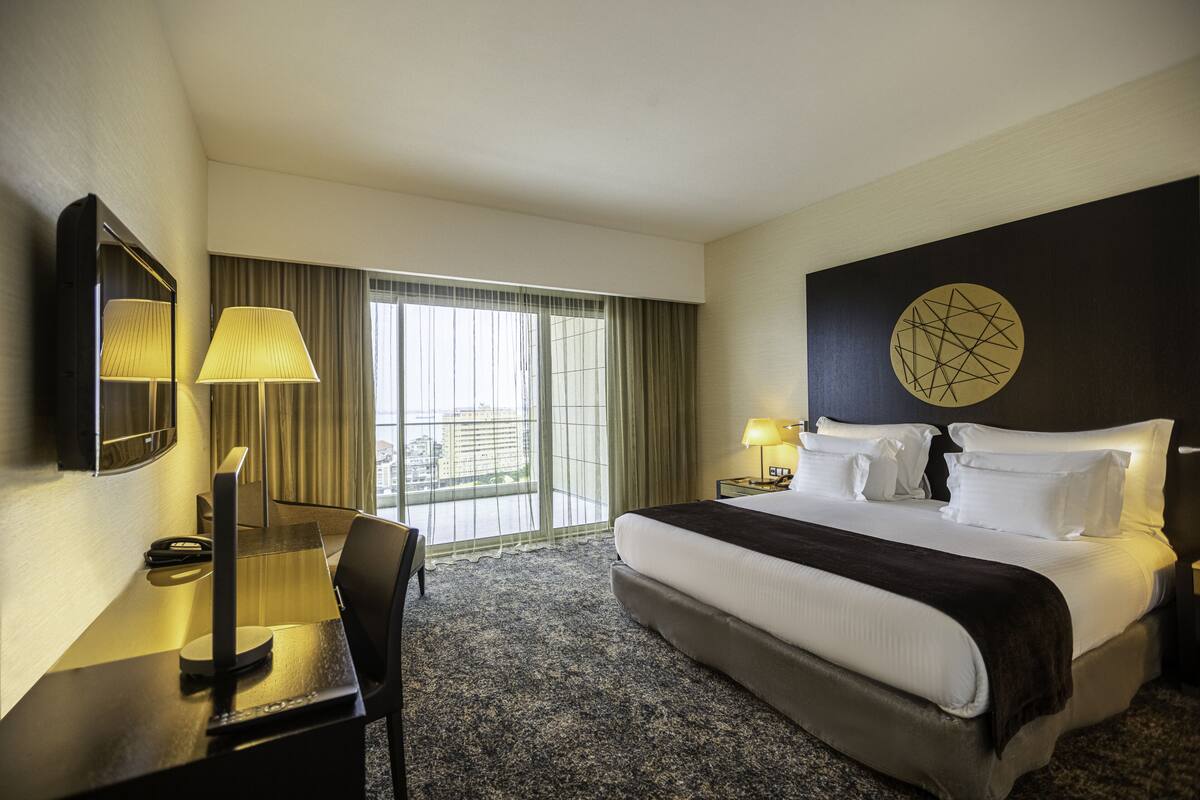A hotel room with a large bed, a desk with a lamp, a chair, and a view of the city through the window.