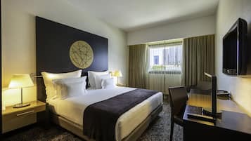 Una habitación de hotel con una cama grande, dos mesitas de noche con lámparas, un escritorio con una computadora y un control remoto, un televisor en la pared y una ventana con cortinas.