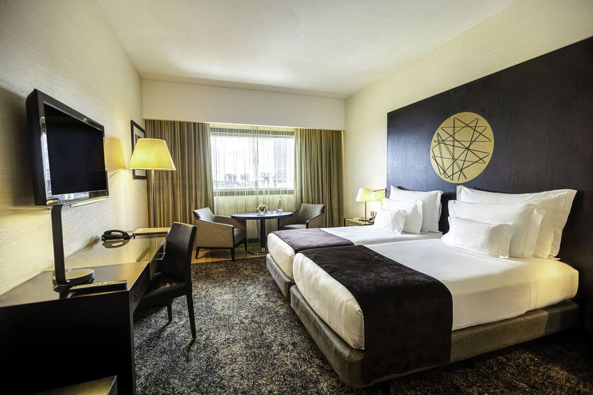 A hotel room with a bed, desk, TV, chair, and a view of the city.