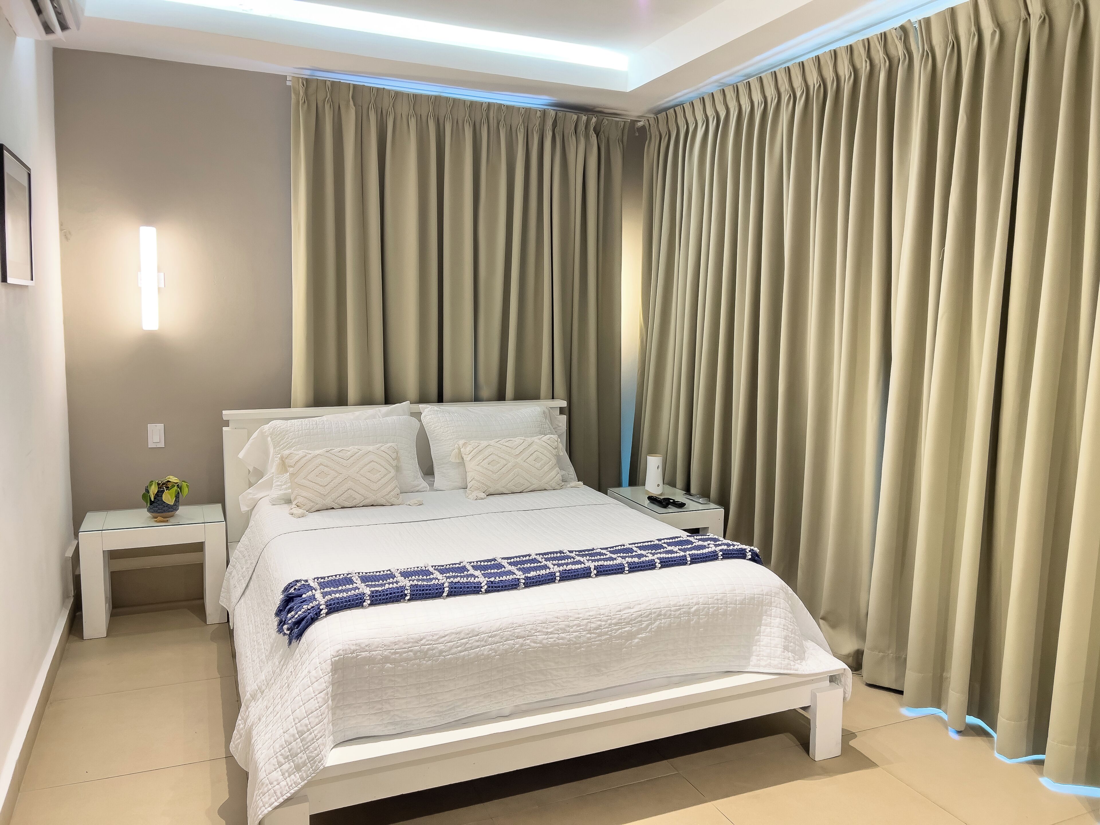 A neatly made bed with white bedding and a blue patterned throw, a bedside table with a lamp and a phone, and a wall-mounted light fixture.