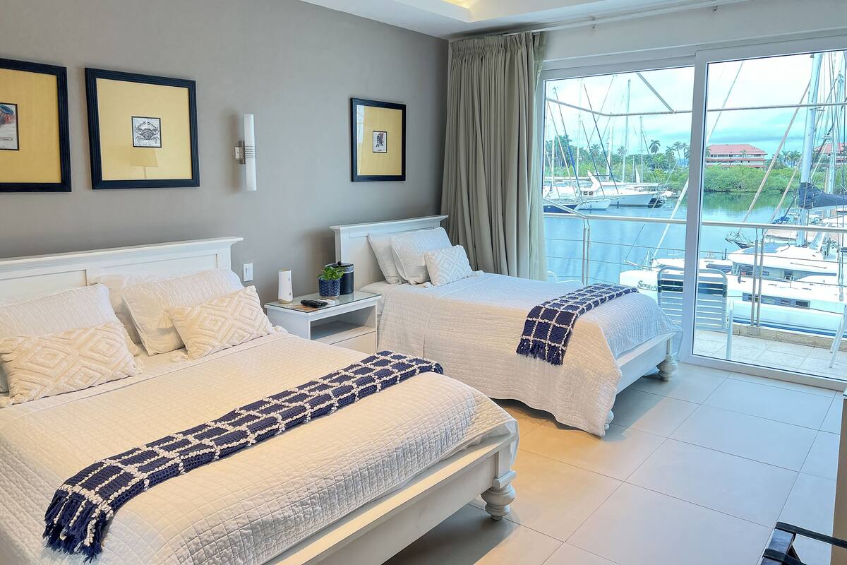 A hotel room with two beds, a large window with a view of boats, and framed pictures on the wall.