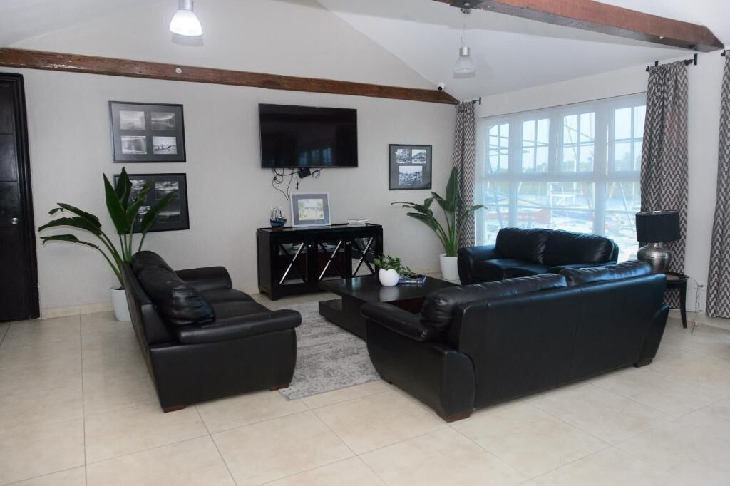 A living room with a black leather sofa, a coffee table, and a TV mounted on the wall. There are framed pictures on the wall and a potted plant.