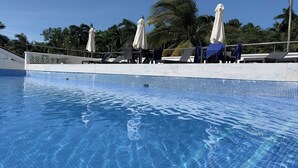 Ein klares, blaues Schwimmbecken mit gefliesten Rändern, Liegestühlen und Sonnenschirmen, umgeben von Palmen.