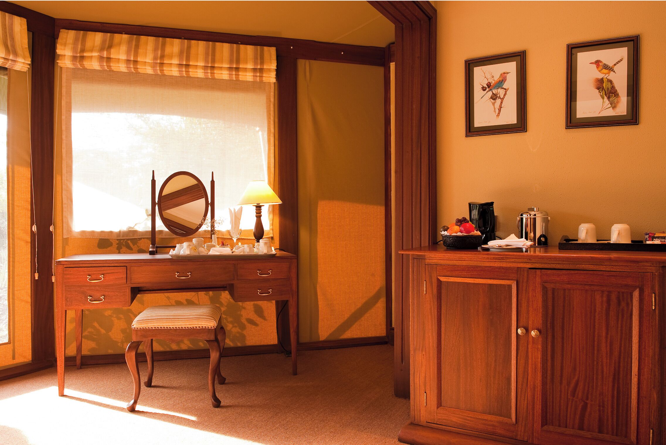 A wooden desk with a mirror, a lamp, and a small bench. There is a cabinet with a tray of fruits and a coffee maker.