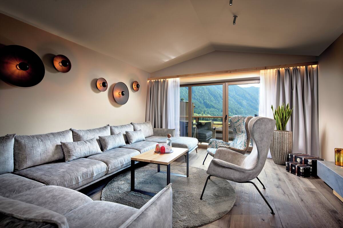 Ein modernes Wohnzimmer mit einem grauen Ecksofa, einem Holztisch und einem großen Fenster mit Vorhängen.