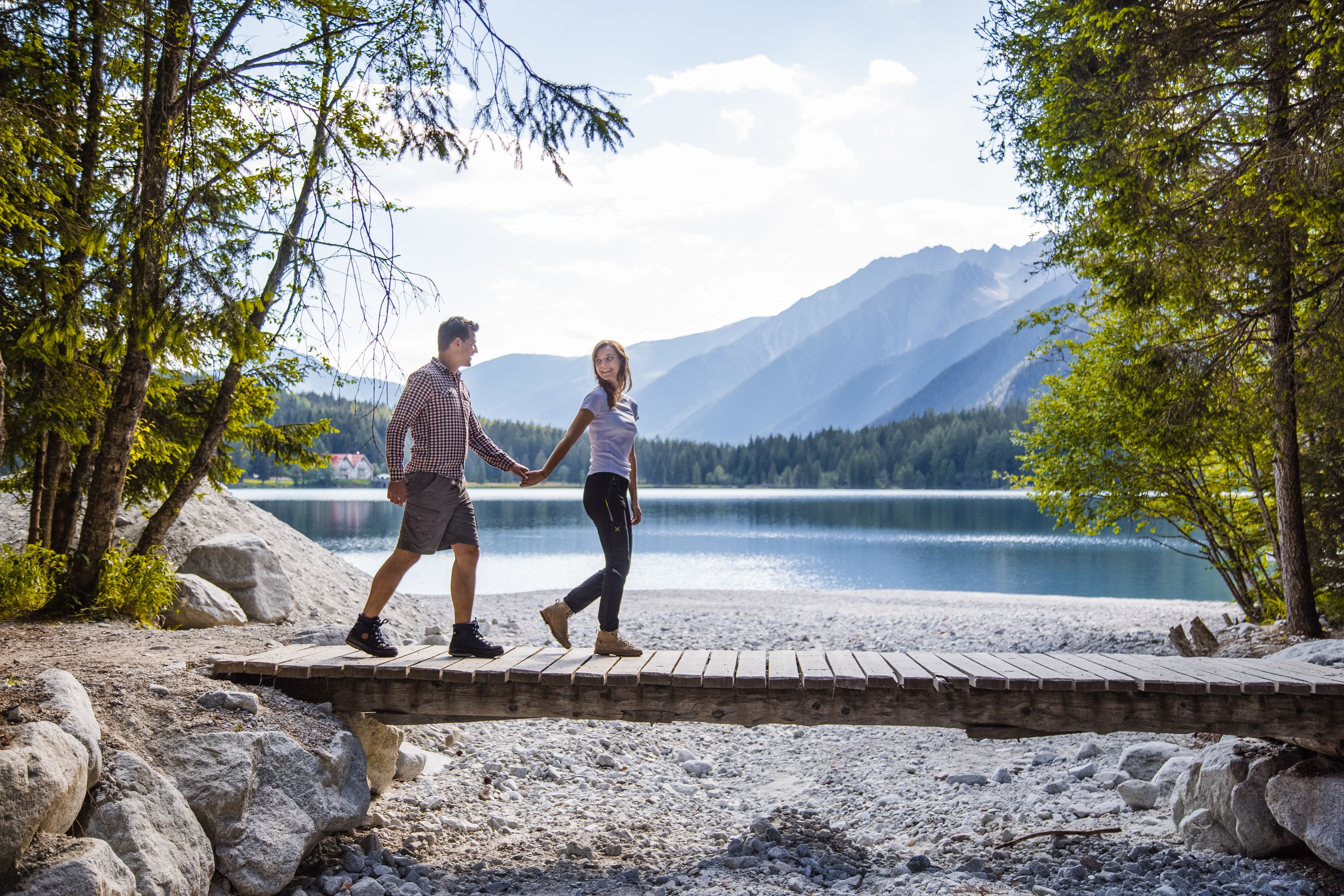 A couple walking on a wooden bridge over a lake with mountains in the background.