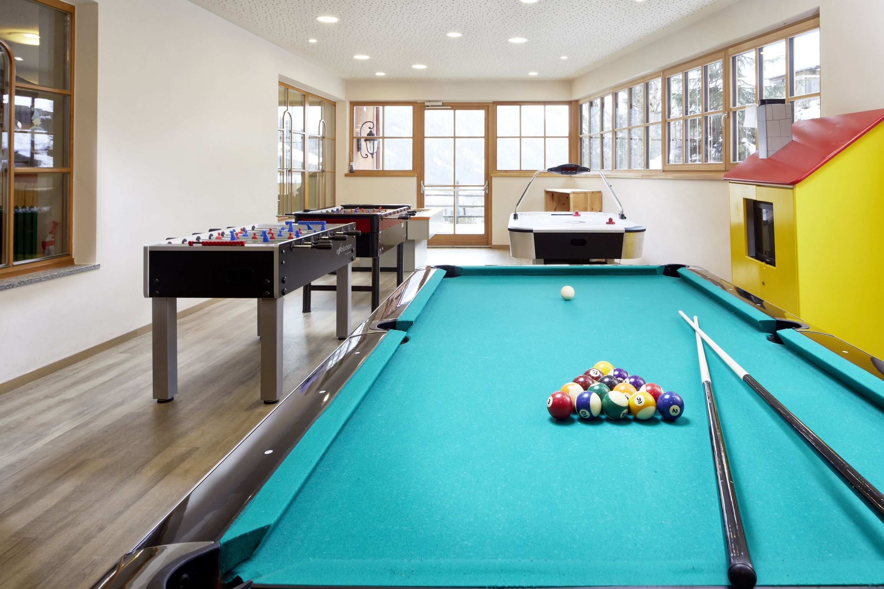 Una mesa de billar con bolas, una mesa de futbolín y una mesa de billares en una habitación con ventanas amplias y vistas al exterior.